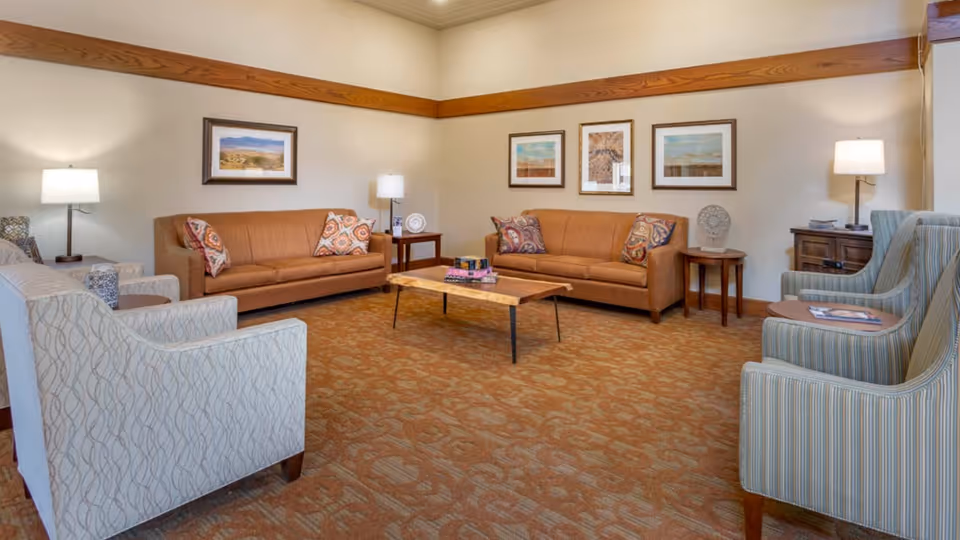 Well-lit communal living room with two sofas, several armchairs, a central coffee table, framed artwork, and lamps.