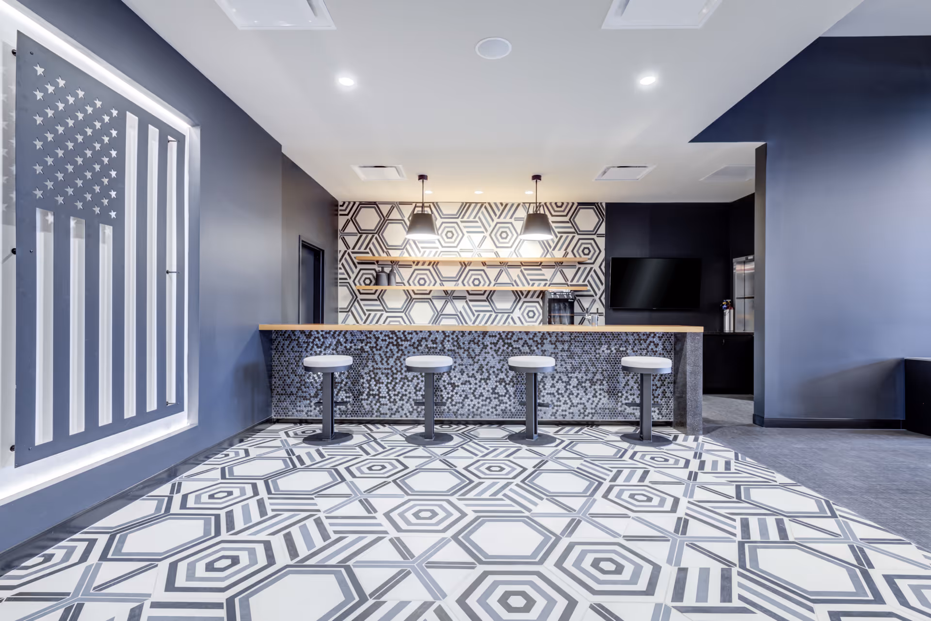 Modern interior space with a patterned tile floor and a bar counter with four stools. The back wall features a geometric hexagonal pattern with two pendant lights hanging above wooden shelves. A large American flag artwork is displayed on the left wall, and a flat-screen TV is mounted on the right wall.