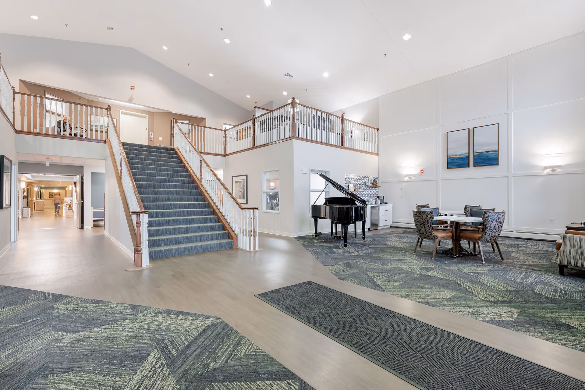 Spacious interior common area of a senior living facility featuring a wide staircase with blue carpeting leading to an upper level, a black grand piano, a round table with four chairs, modern wall art, and soft lighting. The floor has a mix of wood and patterned carpet, and the walls are painted white with wood trim accents.