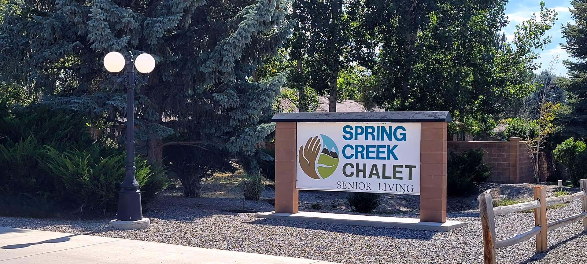 Entry sign reading "Spring Creek Chalet Senior Living" next to a lamppost with trees and landscaping.