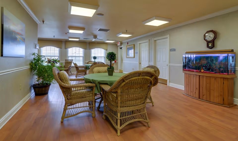 A senior living facility common area with a round table covered by a green tablecloth surrounded by wicker chairs. The room has wooden flooring, several windows with blinds, potted plants, a wall clock, and a large fish tank on a wooden stand. The walls are light-colored with white trim, and there are ceiling lights and a ceiling fan.
