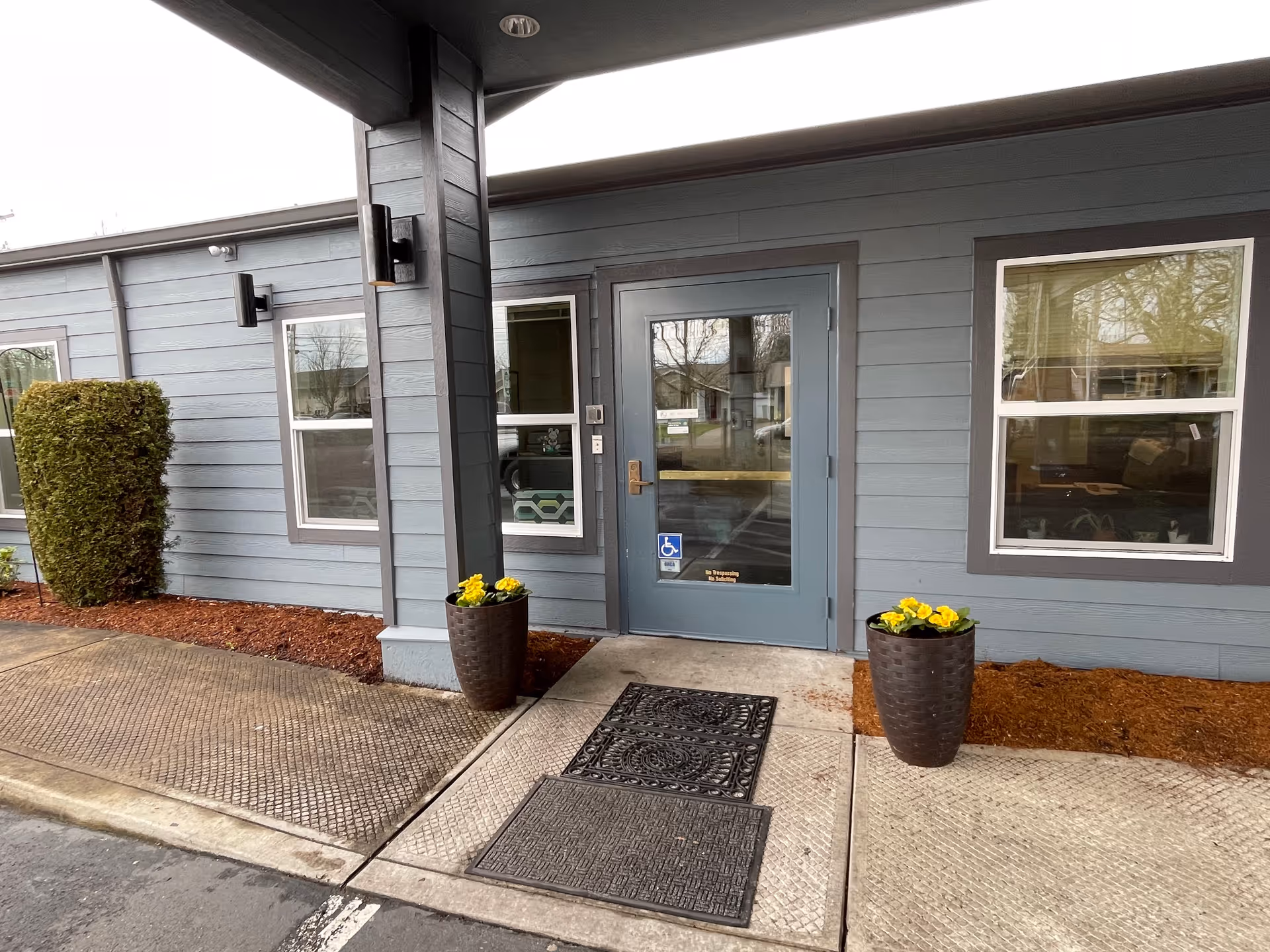 Entrance to a senior living facility with a blue door featuring a handicap accessibility sign. The building exterior is painted gray with two windows on either side of the door. There are two large planters with yellow flowers flanking the entrance, and a neatly trimmed bush to the left. The ground is paved with textured concrete and a black doormat is placed in front of the door.
