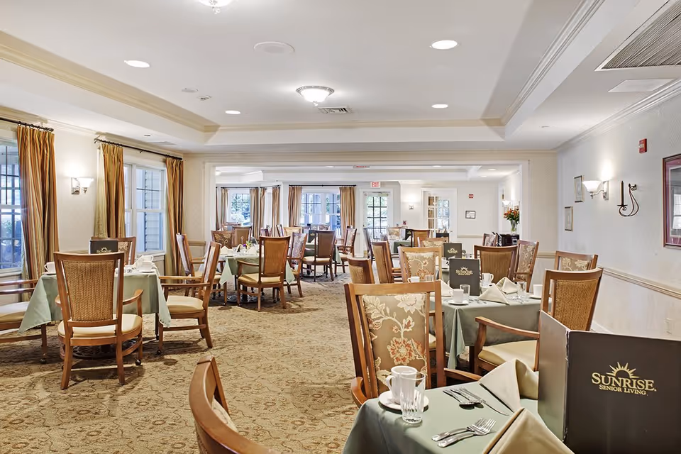 A spacious dining room with multiple tables covered in green tablecloths, set with white napkins, cups, glasses, and silverware. Wooden chairs with cushioned seats and floral upholstery surround the tables. Large windows with brown curtains allow natural light to fill the room. The walls are decorated with framed pictures and sconces, and the carpet has a patterned design.