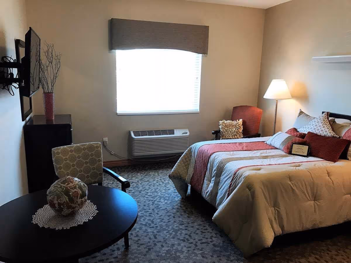 A cozy bedroom in an assisted living facility featuring a bed with a striped comforter and multiple pillows, a red armchair with a decorative pillow, a floor lamp, a round table with a patterned chair and a decorative item on a doily, a wall-mounted TV, and a window with a valance above an air conditioning unit.