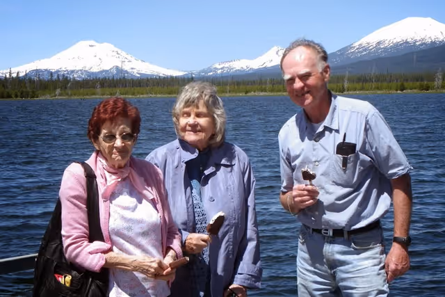 Three elderly people standing outdoors near a large body of water with snow-capped mountains in the background. Two women and one man are smiling and holding ice cream bars. The sky is clear and blue.