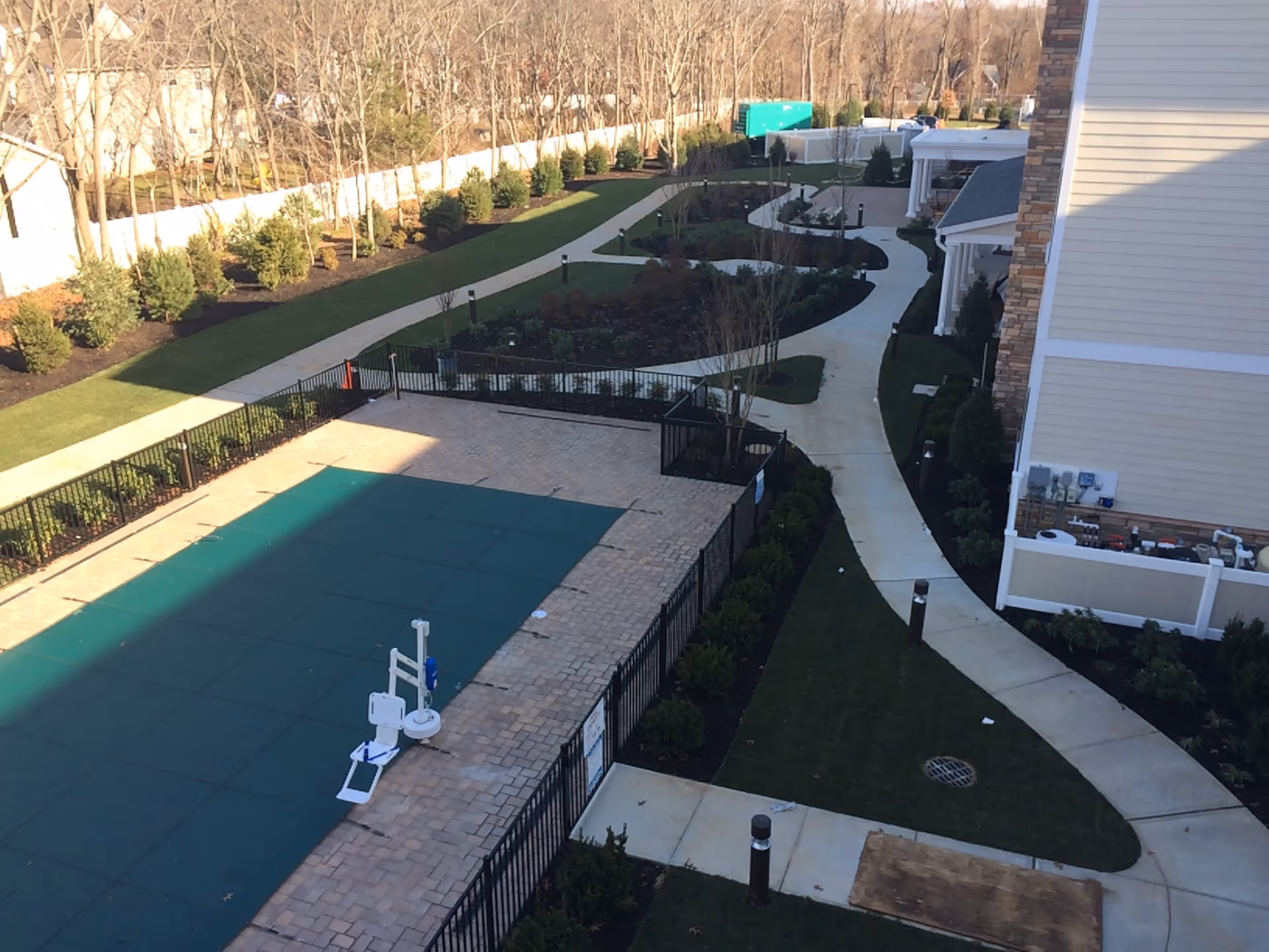 Outdoor view of a senior living facility showing a covered swimming pool surrounded by a black metal fence, paved walkways, landscaped garden areas with bushes and trees, and part of the building exterior with beige siding and stone accents.