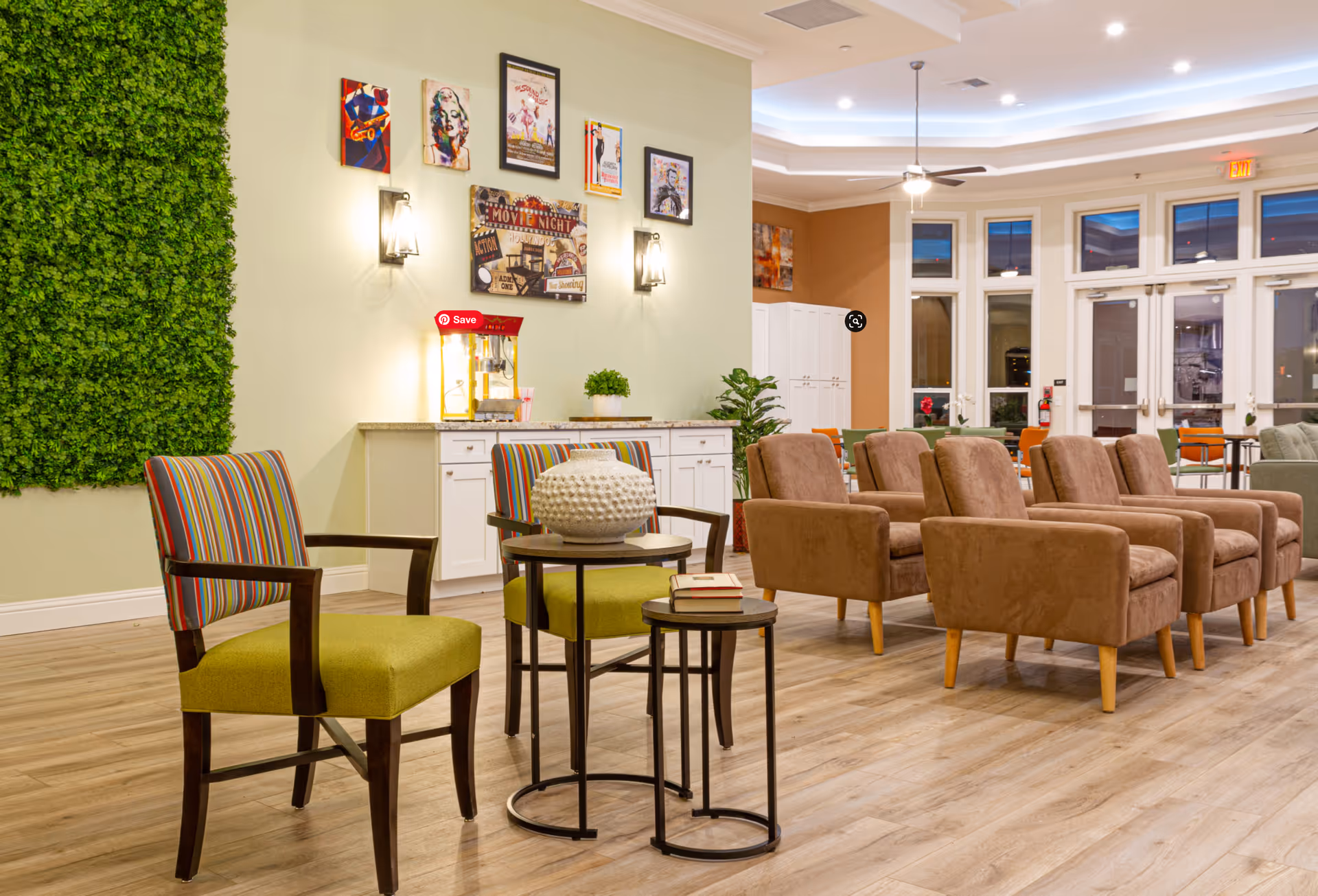 A bright and spacious senior living common area with wooden flooring, featuring a row of brown armchairs, two green chairs with striped backs, and two round nesting tables with decorative items. The wall has framed artwork and sconces, and there is a popcorn machine on a white cabinet. Large windows and glass doors allow natural light to enter the room.