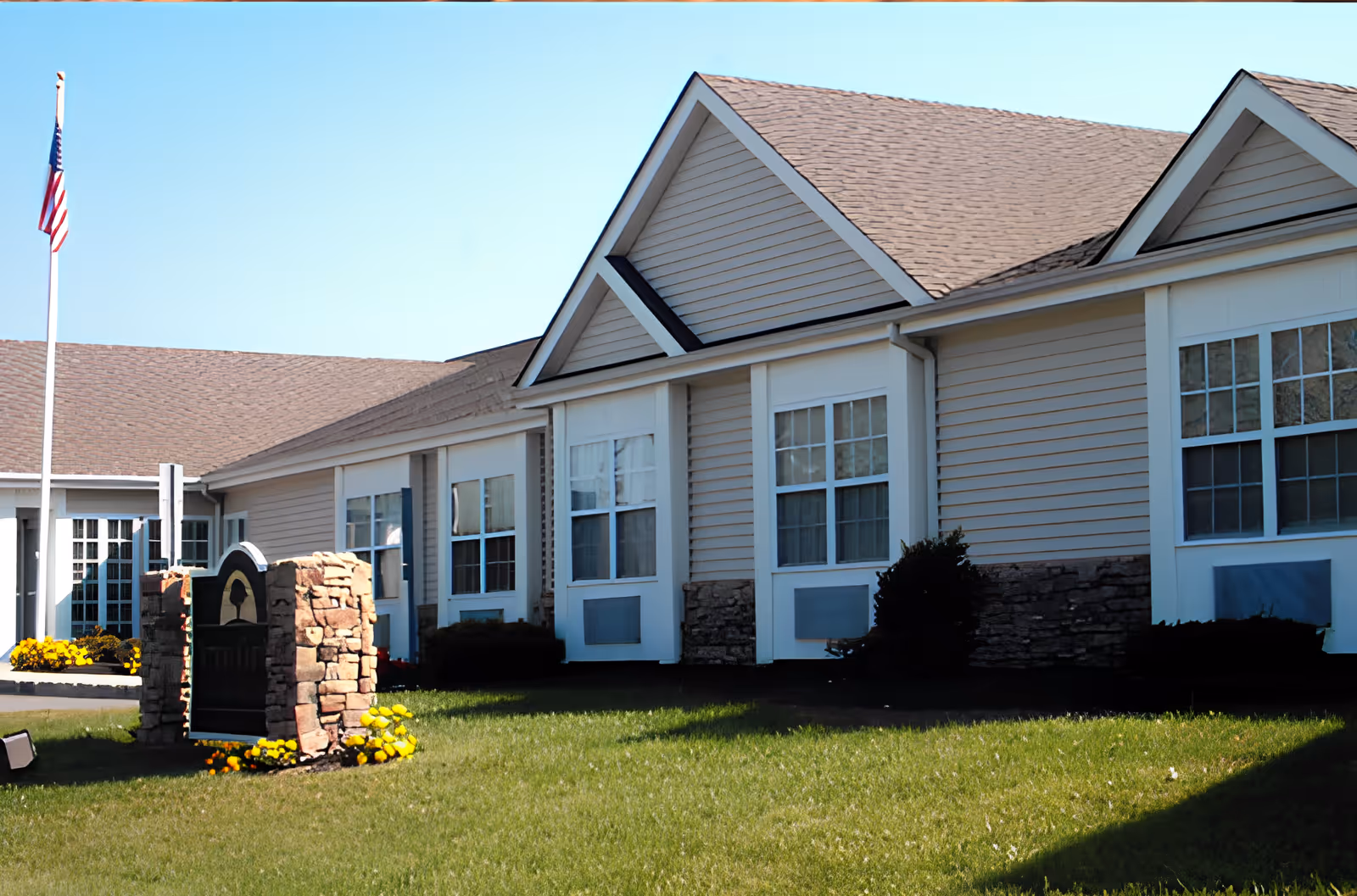 Front exterior of a single-story assisted living building with beige siding, multiple windows, a flagpole, and a landscaped lawn.
