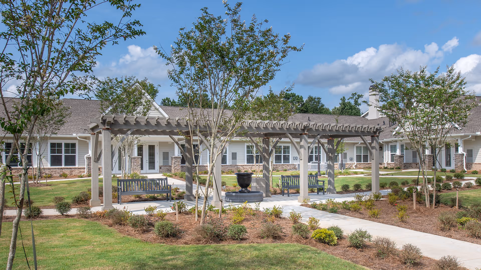 Outdoor courtyard area at Thrive at Green Island featuring a pergola with benches underneath, surrounded by landscaped garden beds, young trees, and a paved walkway. The background shows a single-story building with white siding and brick accents under a blue sky with scattered clouds.