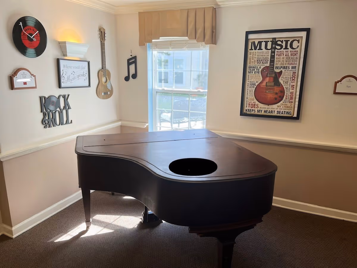 A music-themed room with a closed grand piano in the center. The walls are decorated with a vinyl record clock, a small acoustic guitar, a musical note wall hanging, a framed quote saying 'Where words fail music speaks', a 'Rock & Roll' wall decoration, and a large framed poster featuring a guitar and various music-related words. There is a window with blinds letting in natural light.