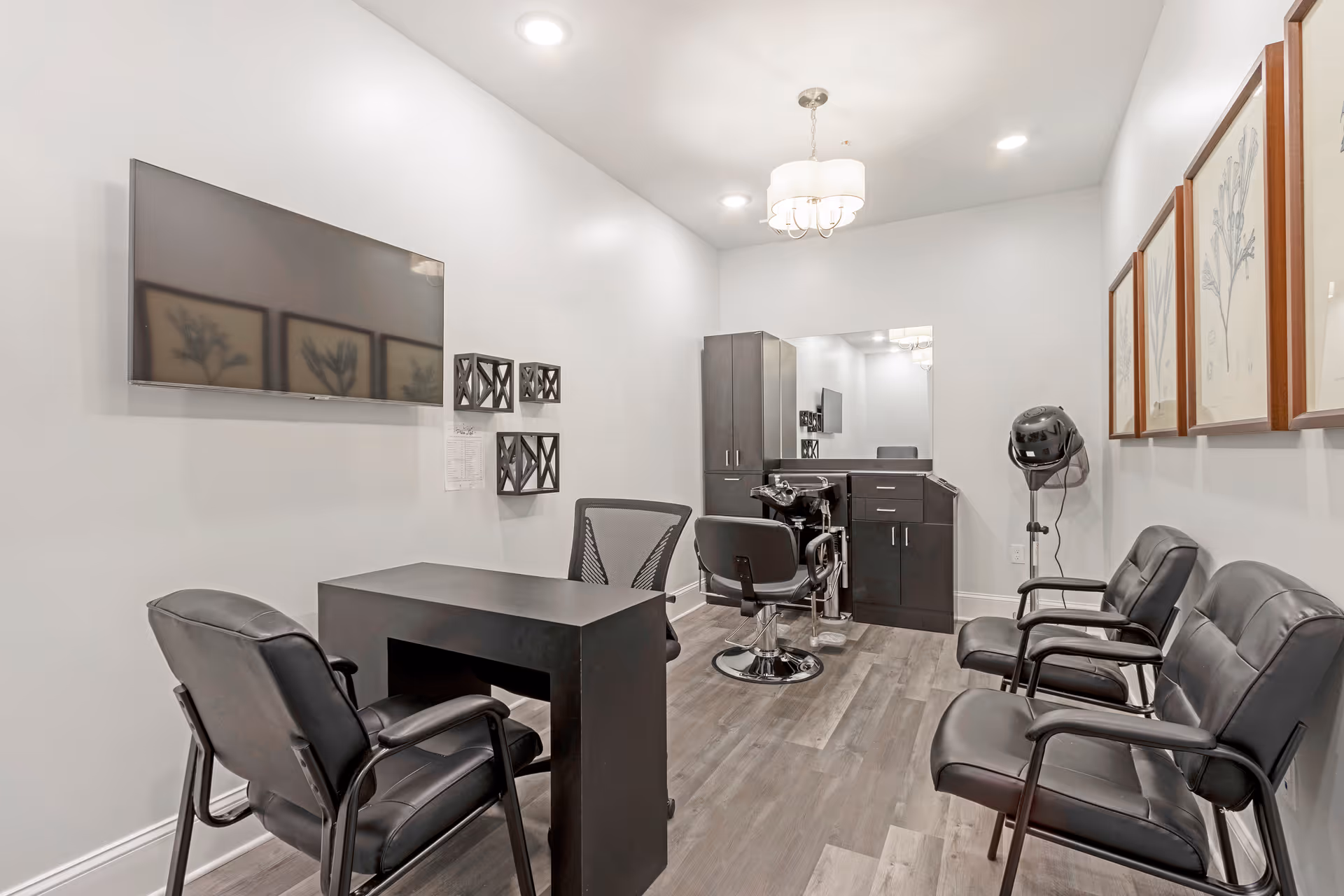A modern salon room with a black styling chair in front of a large mirror and cabinetry. There is a hair dryer hood on a stand, a black desk with a mesh office chair, and three black leather waiting chairs along the wall. The room has light gray walls, wood flooring, framed botanical artwork, and a wall-mounted flat screen TV.