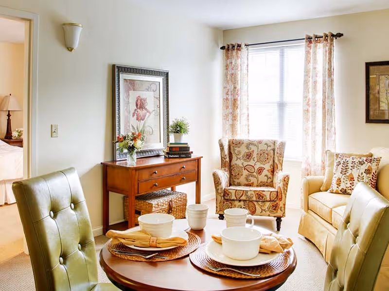 A cozy living room area with a round wooden table set for four with white bowls, cups, and yellow napkins. The room features two green cushioned chairs around the table, a floral patterned armchair near a window with floral curtains, and a beige sofa with decorative pillows. A wooden console table against the wall holds a framed floral artwork, a vase with flowers, and a small plant. An adjacent bedroom is partially visible through an open door.