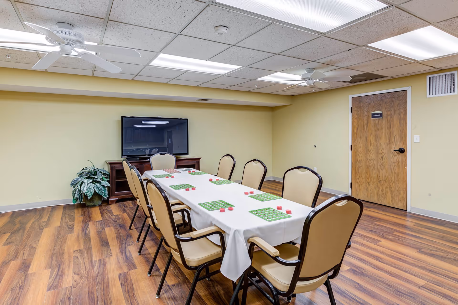 A room with a long table covered with a white tablecloth and eight beige cushioned chairs around it. On the table are green bingo cards and red bingo chips. The room has wood flooring, pale yellow walls, a wooden door labeled 'Storage', a flat-screen TV on a wooden stand, two ceiling fans, and fluorescent lighting.
