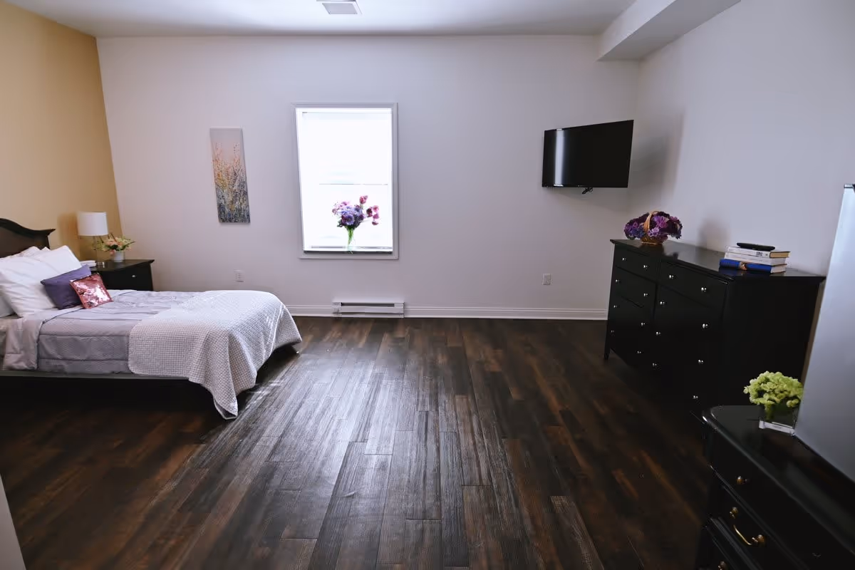 A bedroom with a bed on the left side featuring white and purple bedding, a nightstand with a lamp and flowers, a window with a vase of purple flowers on the sill, a wall-mounted TV on the right wall, and a black dresser with books and a flower arrangement. The room has dark wooden flooring and light-colored walls.