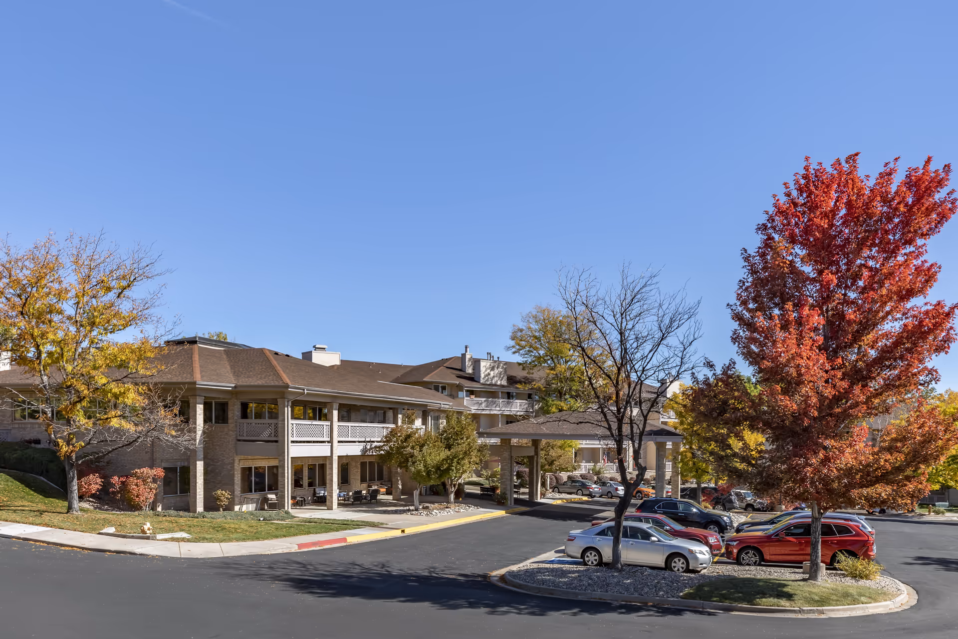 Front exterior of a two-story senior living building with a covered entrance, parked cars, and autumn trees.