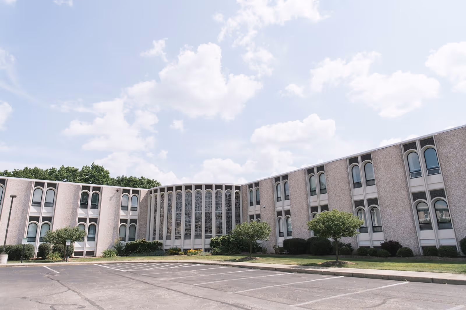 Exterior view of a two-story building with multiple arched windows and a central section featuring tall vertical windows with decorative patterns. The building is surrounded by small trees and bushes, with an empty parking lot in the foreground under a partly cloudy sky.