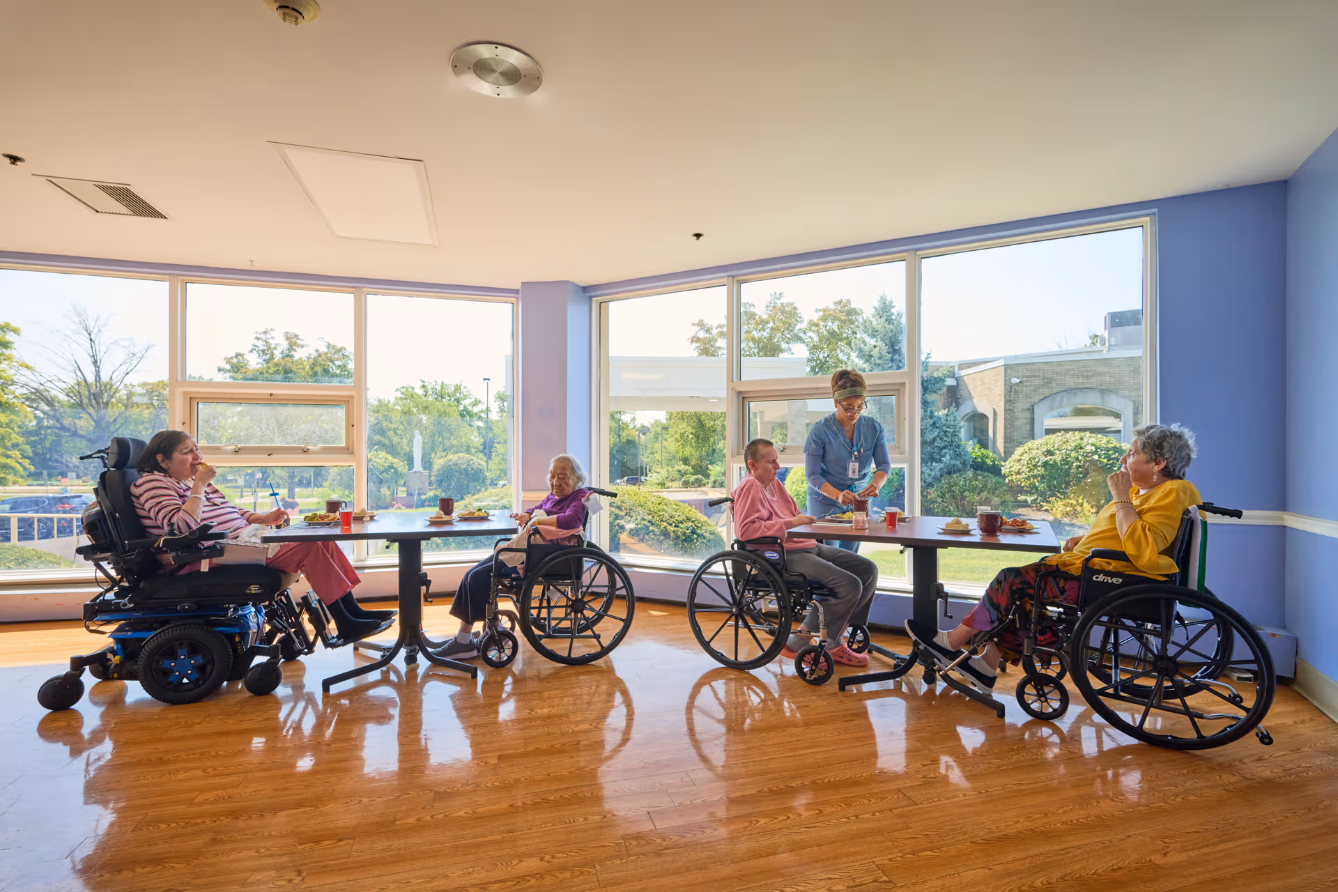 Four elderly residents in wheelchairs sit around tables in a sunny dining room with large windows while a caregiver assists them.