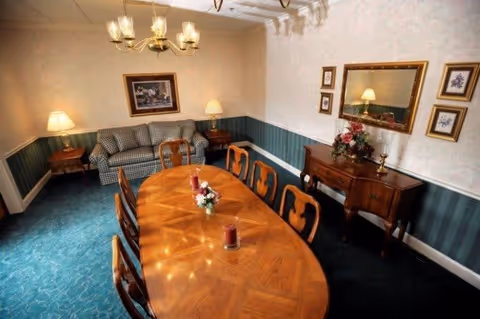 A traditional dining room with a long wooden dining table surrounded by eight chairs. The table has decorative candles and a floral centerpiece. Against the wall is a wooden sideboard with a flower arrangement and a large mirror above it. There is a patterned sofa and two table lamps on either side of the room. The walls have a two-tone design with wallpaper above and green striped paneling below, and the floor is carpeted in blue.