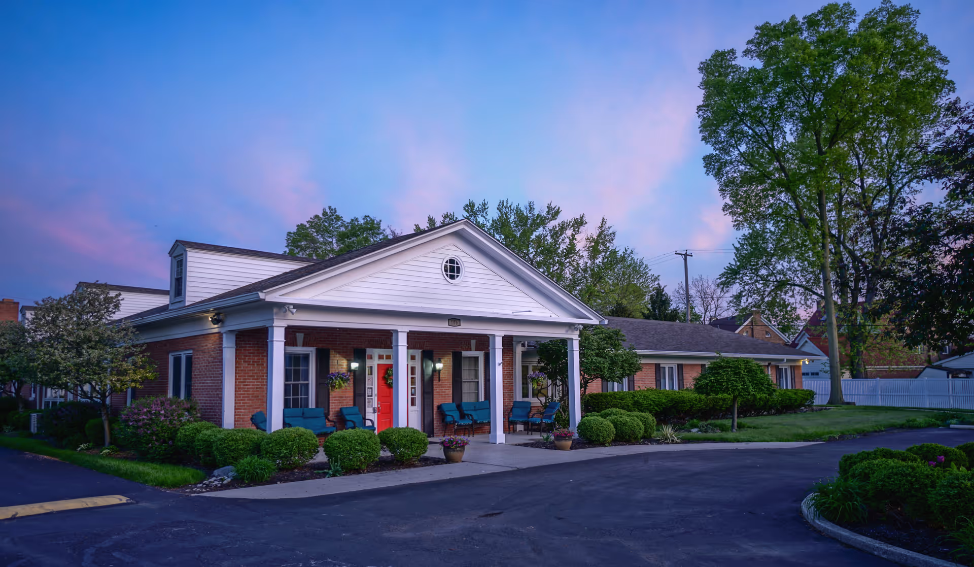 Exterior view of a single-story brick building with white columns and a red door, surrounded by well-maintained bushes and trees under a purple and blue evening sky.