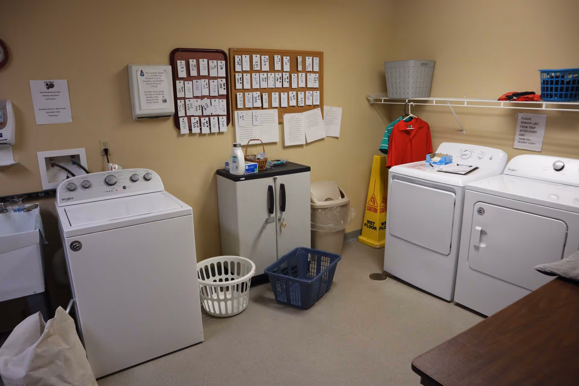 Laundry room with a washing machine and dryer, laundry baskets, a small cabinet with detergent, a trash can, hanging clothes, and bulletin boards with room assignments on a beige wall.