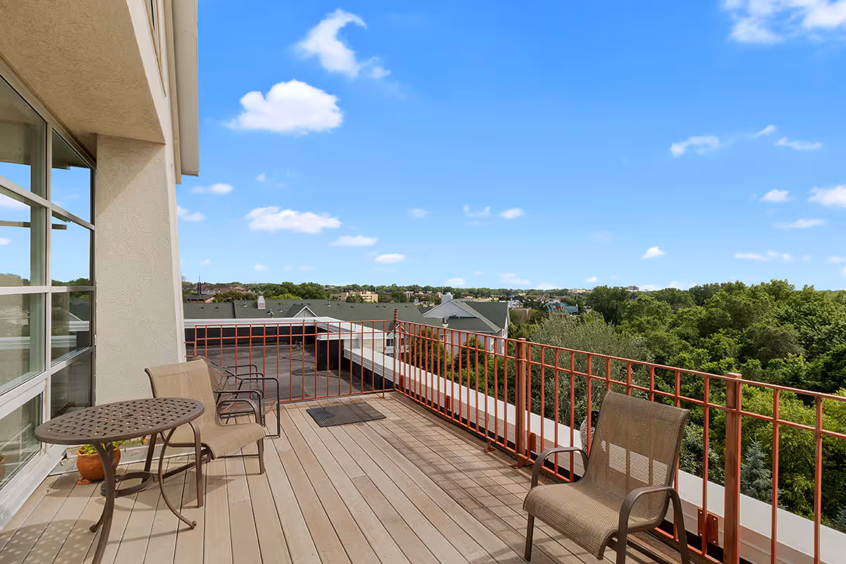 Outdoor balcony area with a metal table and three chairs, overlooking a view of rooftops and green trees under a blue sky with scattered clouds.