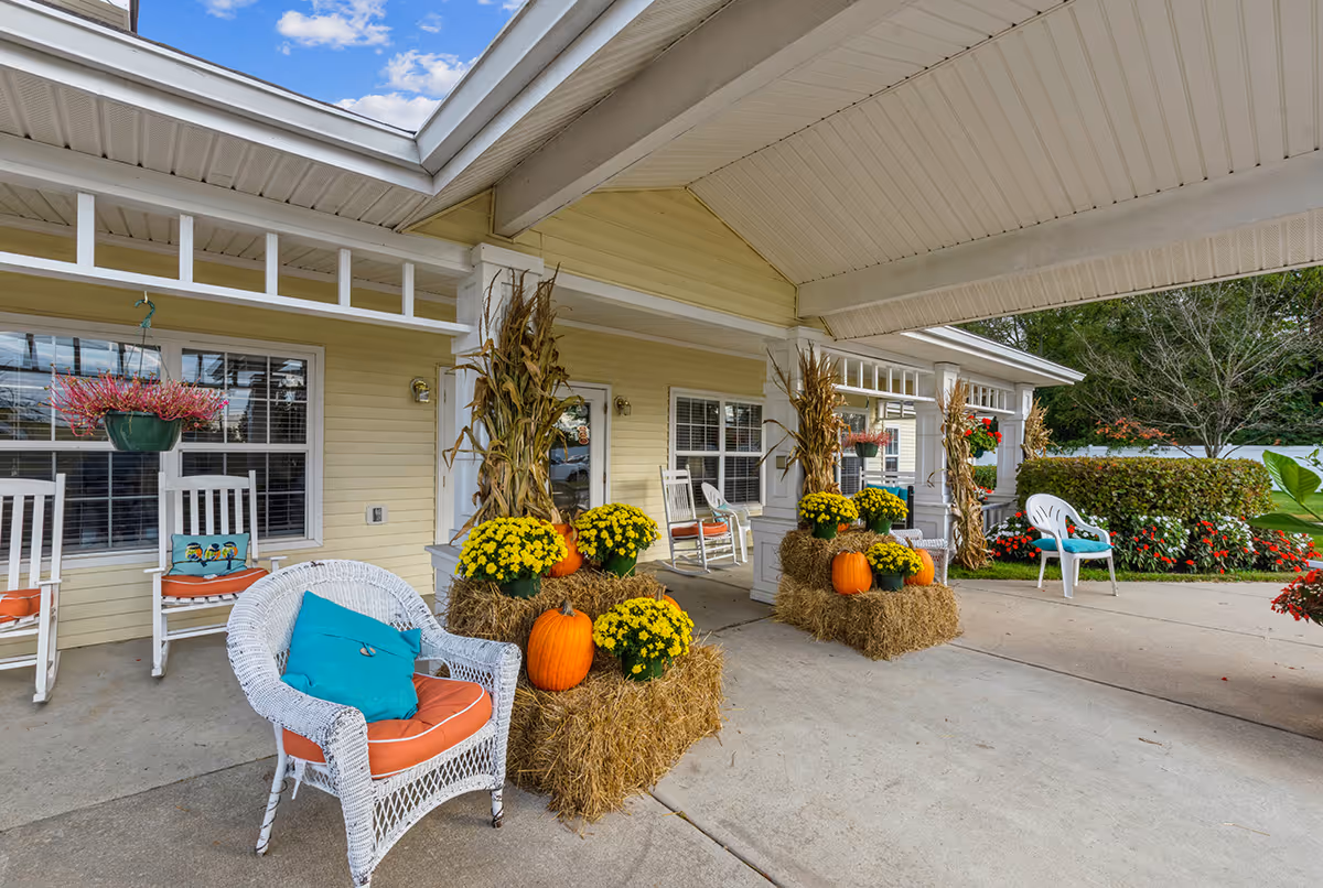 Covered outdoor patio area with white wicker and wooden chairs, decorated with orange pumpkins, yellow flowers, hay bales, and dried corn stalks. The building exterior is light yellow with white trim, and there are hanging flower pots and a garden with green bushes and red flowers in the background.