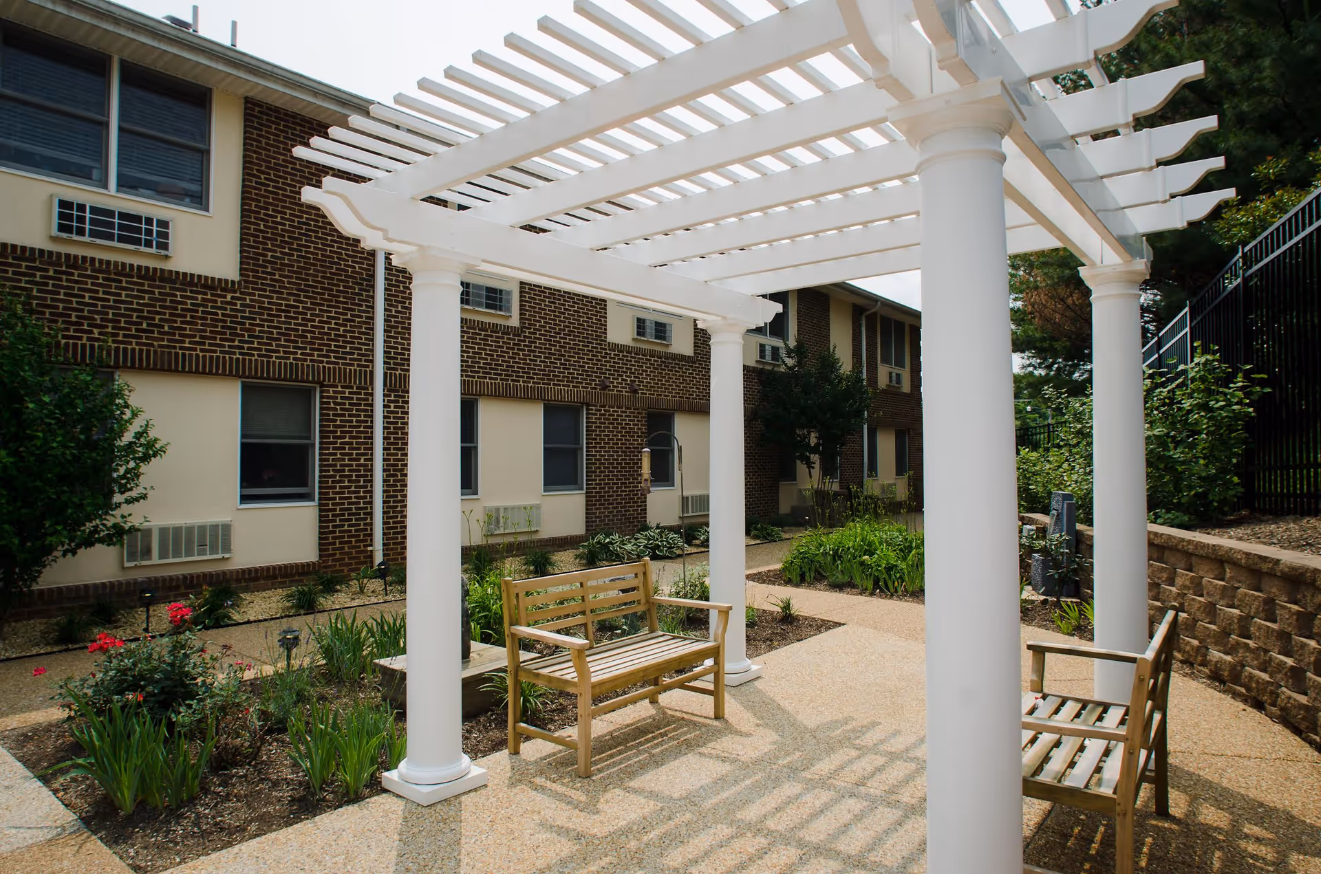 Outdoor seating area with two wooden benches under a white pergola, surrounded by garden beds with plants and flowers, adjacent to a brick building with windows and air conditioning units.