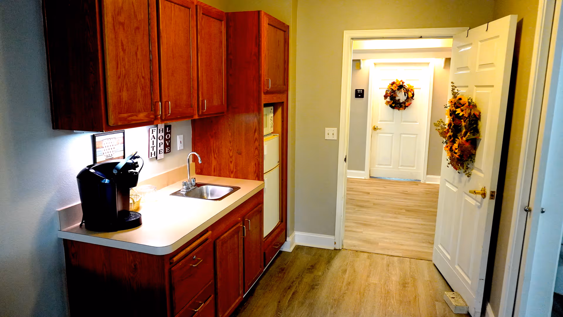 Small kitchenette with wooden cabinets, a sink and coffee maker opening onto a hallway with a door decorated with wreaths.