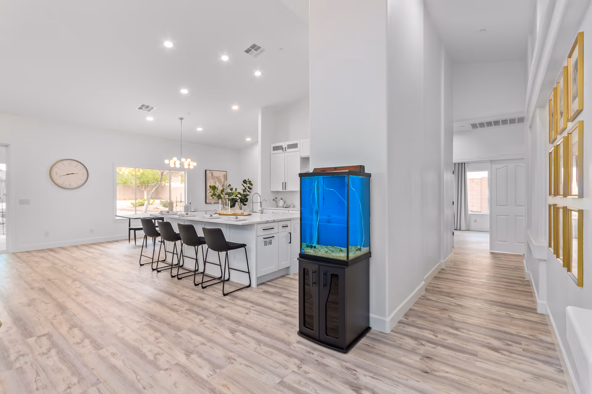 Bright open-plan kitchen and dining area with a large island and bar stools, an aquarium near a hallway, and light wood flooring.