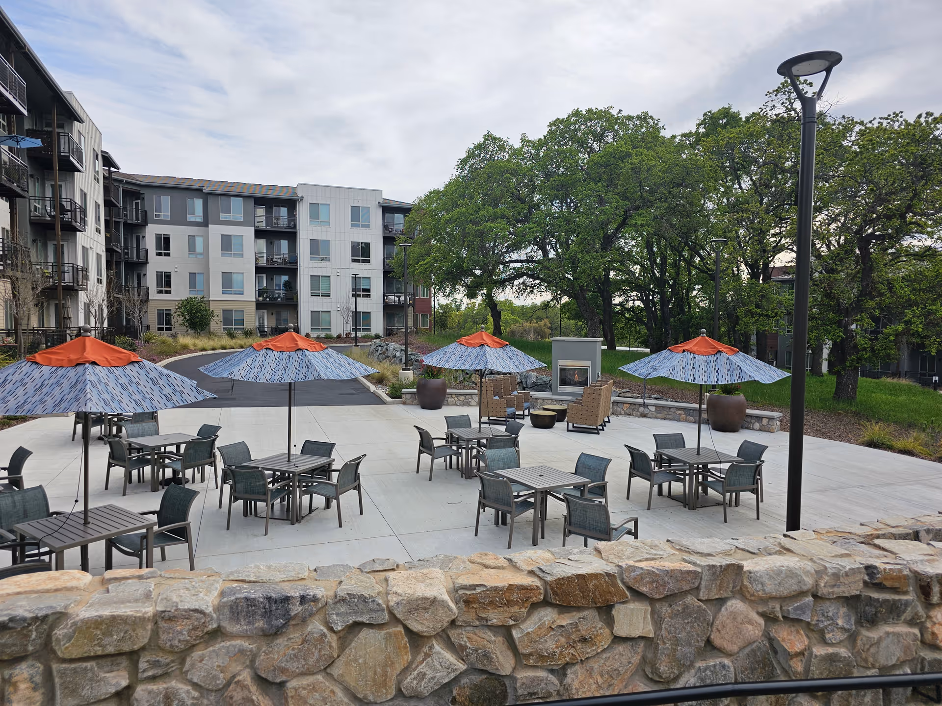 Outdoor courtyard patio with tables, chairs, and blue-orange umbrellas in front of a multi-story senior living building and trees.