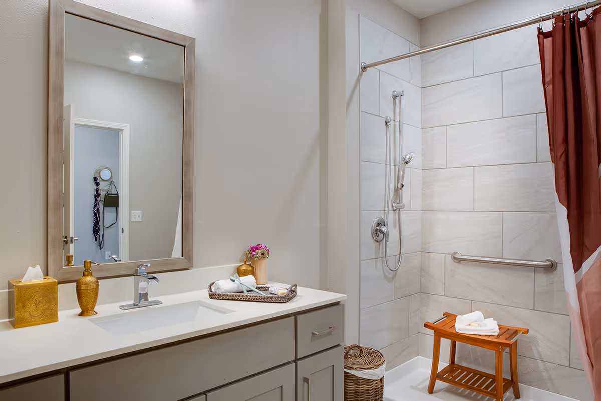A modern bathroom featuring a large mirror above a white countertop with a sink. On the countertop are a gold tissue box, a gold soap dispenser, and a tray with towels and a small vase of flowers. The shower area has light beige tiles, a handheld showerhead, a grab bar, and a wooden shower bench with folded towels on it. A red and white shower curtain is partially visible.