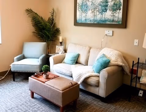 Cozy sitting area with a loveseat, armchair, ottoman, decorative pillows, a potted plant and wall art in a living room space.