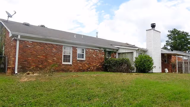 Single-story red brick residential building with a white chimney and a grassy front lawn under a partly cloudy sky.