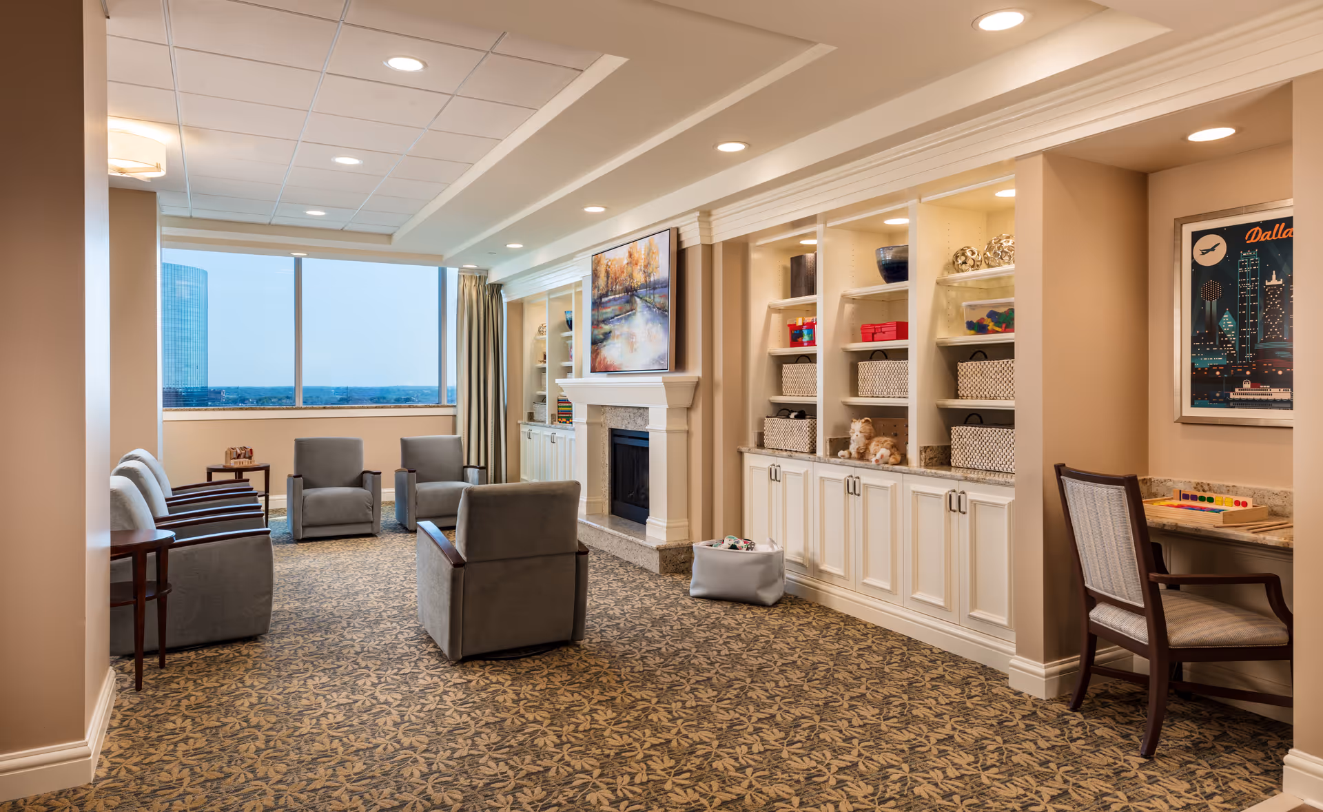 A cozy senior living common area with several gray armchairs arranged around a fireplace with a painting above it. Built-in shelves with baskets and decorative items line one wall. A large window offers a view of a cityscape with a tall building. A small desk with a chair and a colorful game is positioned in a corner.