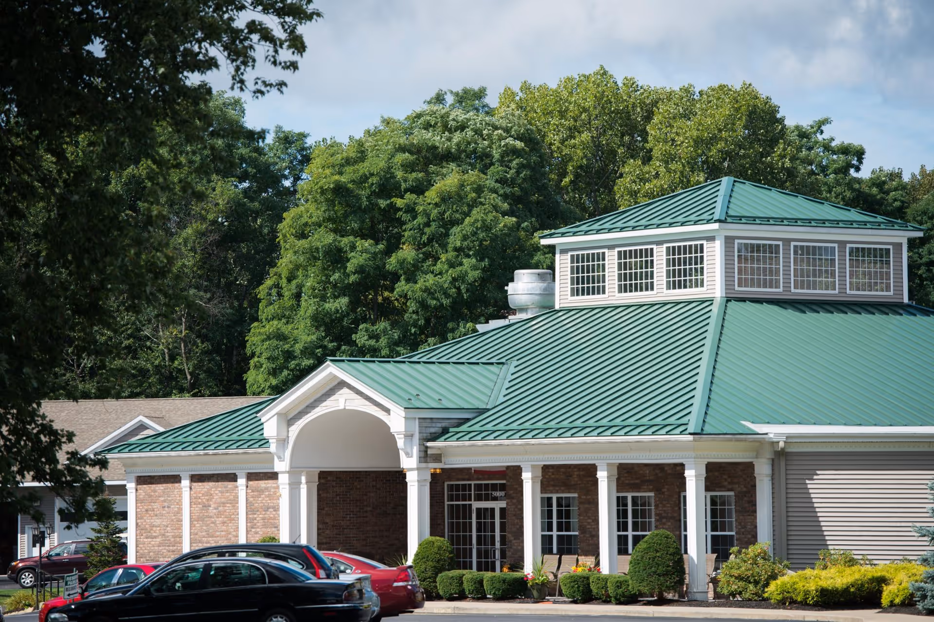 Exterior view of a building with a green metal roof and brick walls, surrounded by trees and parked cars in front. The building has white columns and large windows, with a small garden area featuring shrubs and flowers near the entrance.