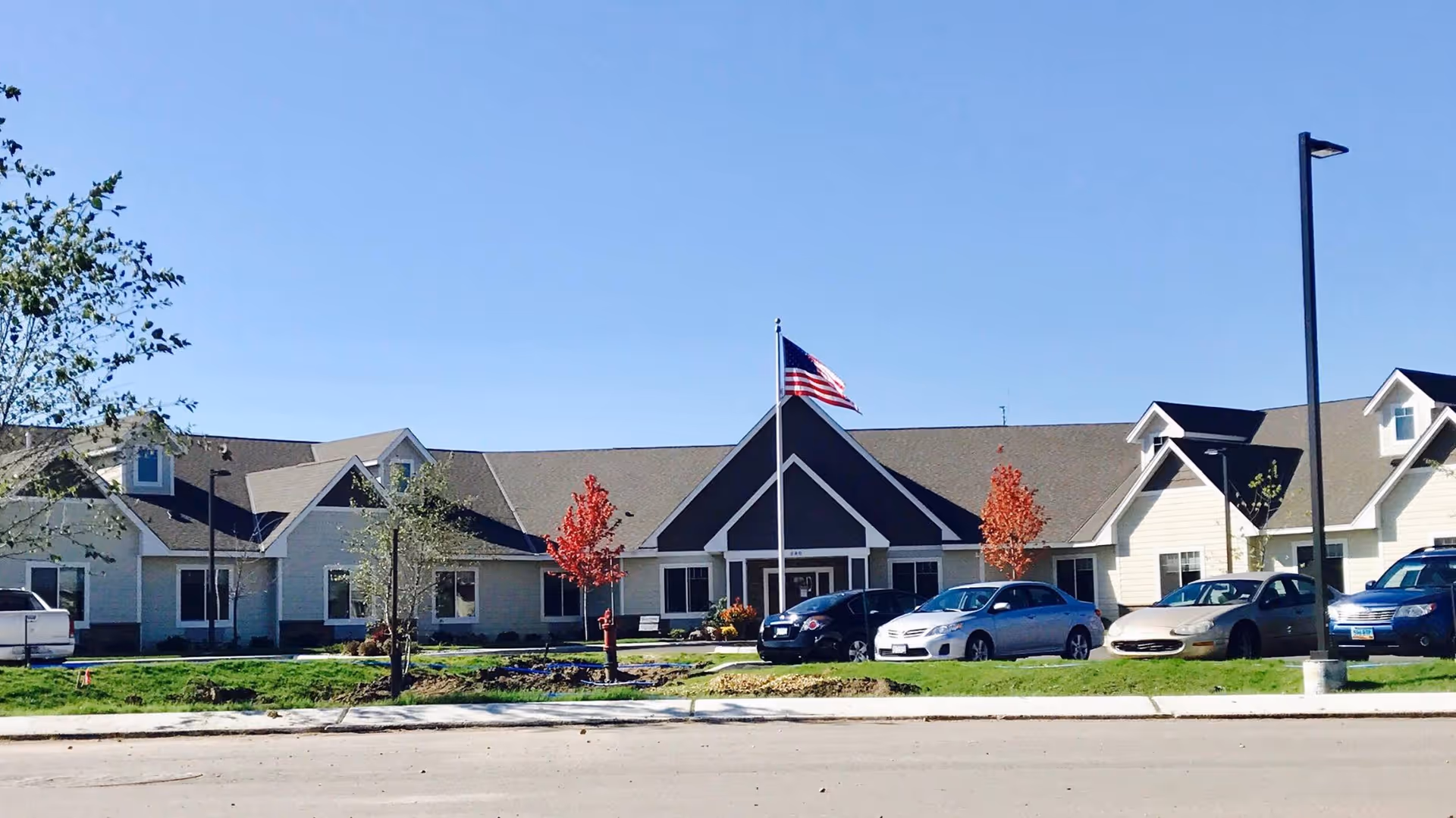 Exterior view of a single-story assisted living facility with a peaked roof and multiple windows. There is an American flag on a flagpole in front of the building, several small trees with some red foliage, a sidewalk, and a parking area with several parked cars under a clear blue sky.