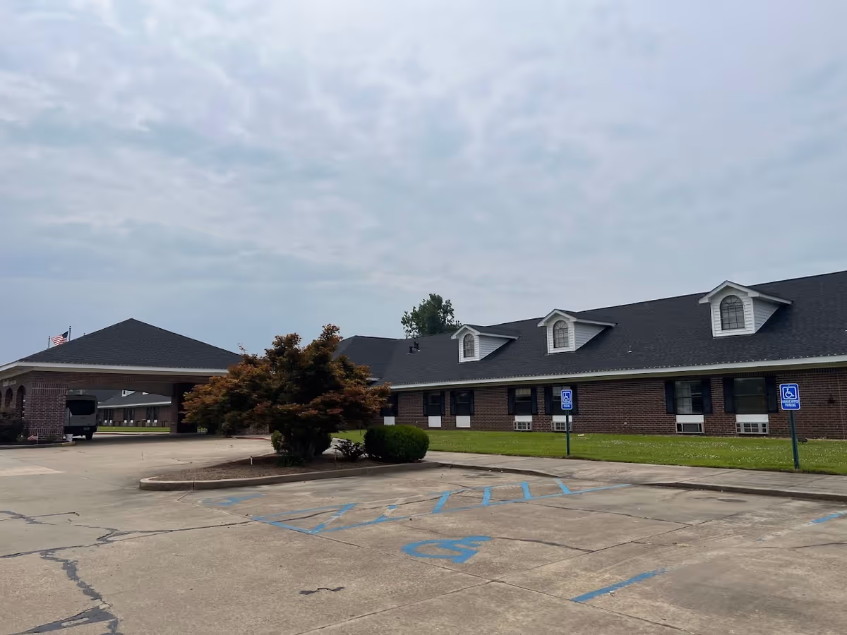 Brick single-story senior living building with dormer windows, a covered entrance, a small landscaped island, and empty handicapped parking spaces under a cloudy sky.