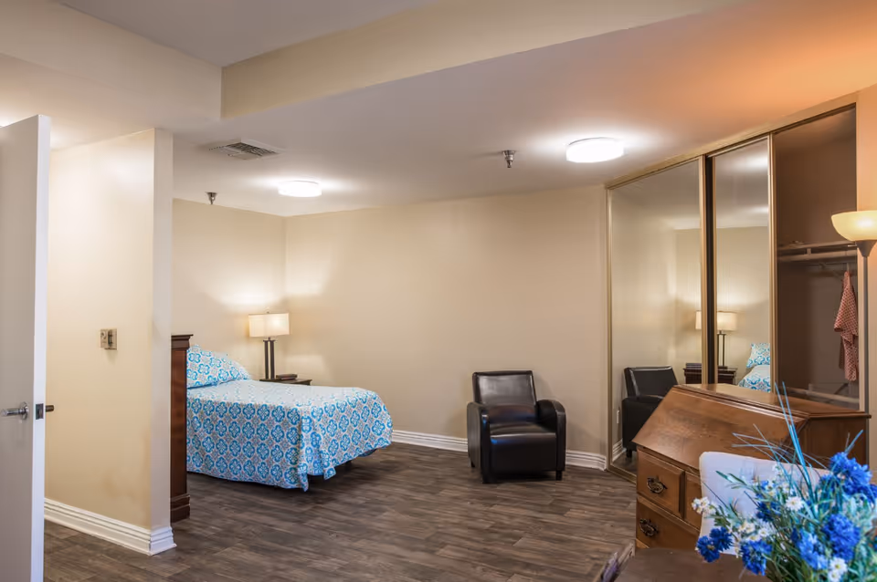 A bedroom in an assisted living facility featuring a bed with blue patterned bedding, a nightstand with a lamp, a black armchair, a wooden dresser, a mirrored closet, and a bouquet of blue flowers on a table in the foreground.