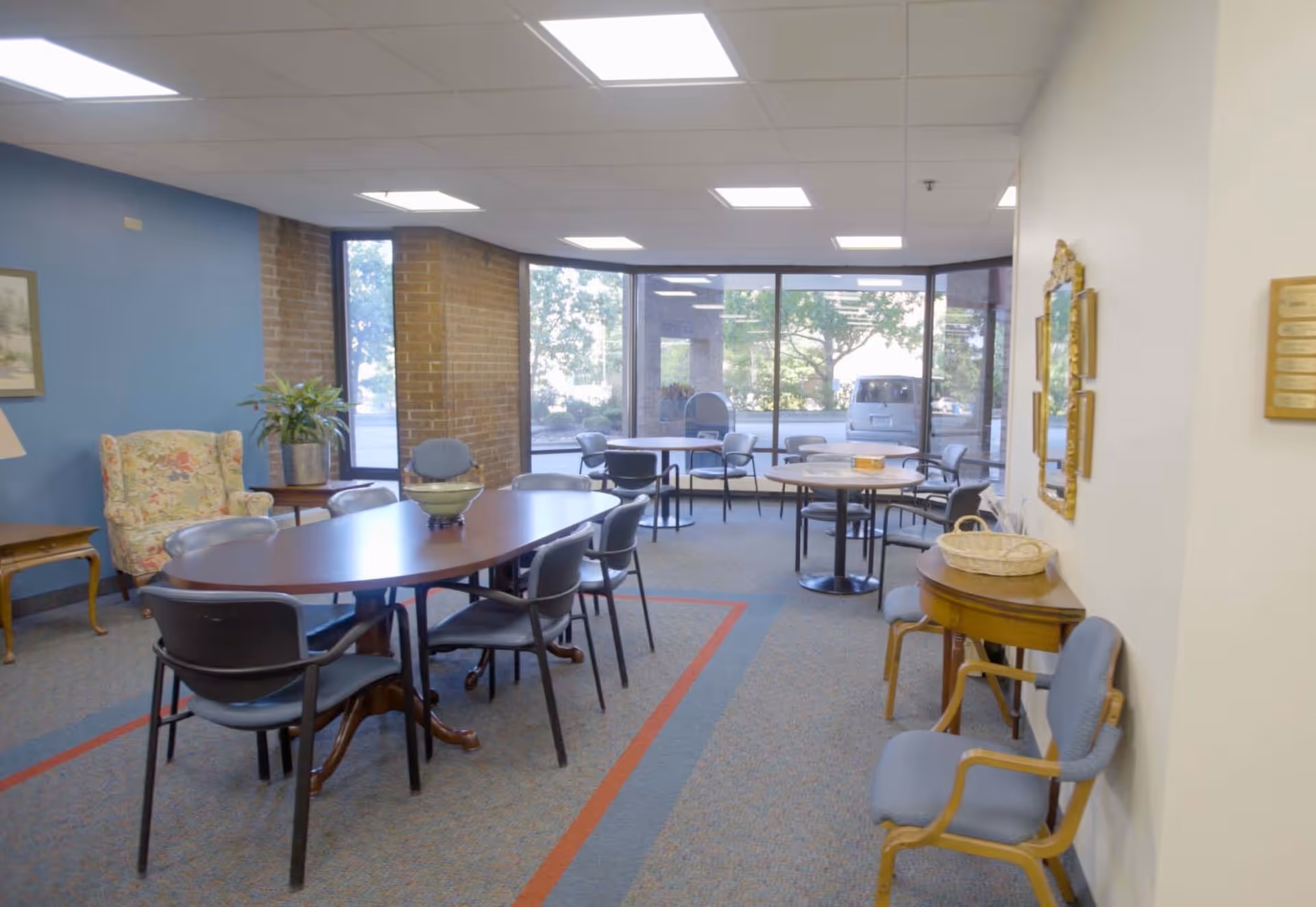 A well-lit common area in a senior living facility with multiple tables and chairs arranged for group seating. The room has large windows letting in natural light, a floral armchair, a potted plant, and a small side table. The walls are painted blue and white, and there is a mirror and a plaque on the wall.