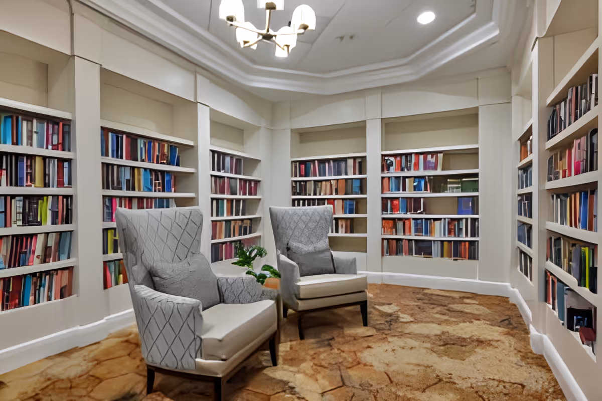 A cozy library room with built-in bookshelves filled with colorful books lining the walls. Two upholstered armchairs with patterned fabric and cushions are positioned in the center on a brown and beige patterned carpet. A modern chandelier hangs from the ceiling, and a small green plant is placed between the chairs.