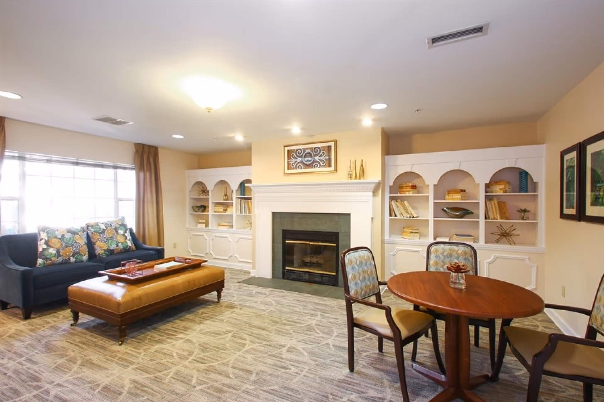A cozy senior living community common area with a dark blue sofa adorned with floral cushions, a large brown leather ottoman, a round wooden table with four cushioned chairs, built-in white bookshelves filled with books and decorative items, and a fireplace with a framed artwork above it. The room is well-lit with ceiling lights and natural light from a large window with brown curtains.