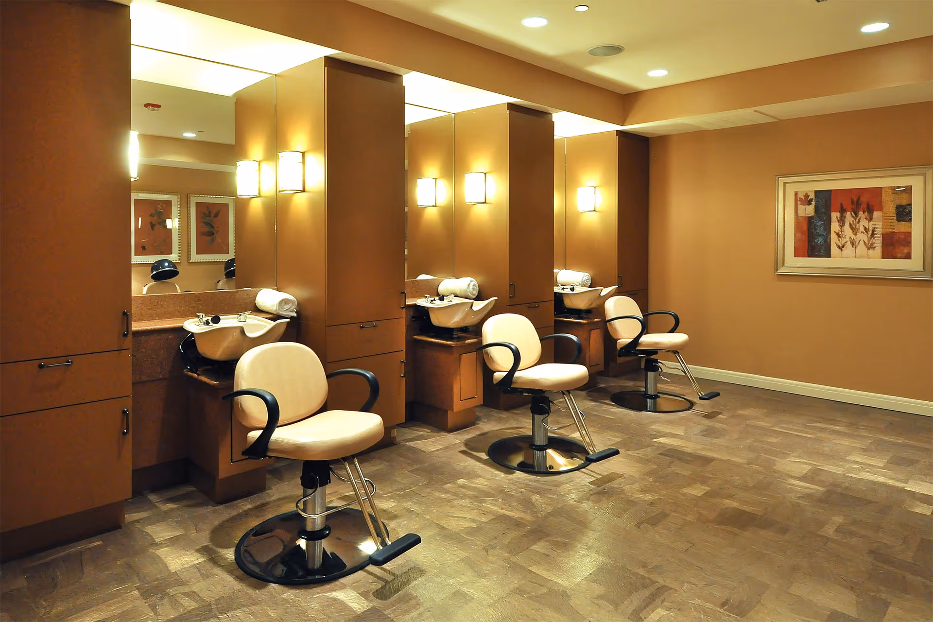 Interior view of a salon area with three beige salon chairs in front of sinks and mirrors, warm lighting, brown walls, and framed artwork on the wall.