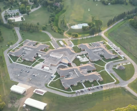 Aerial view of the North Dakota Veterans Home facility showing multiple connected buildings with parking lots, surrounding green lawns, trees, and a small pond in the background.