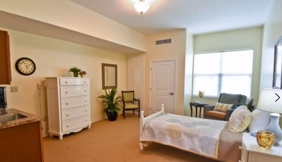 A bright and cozy bedroom in a senior living facility with a single bed covered in a patterned quilt, a white dresser with plants on top, a chair, a small table with a lamp and decorative items, a large window with blinds, and a kitchenette area with a sink and countertop.