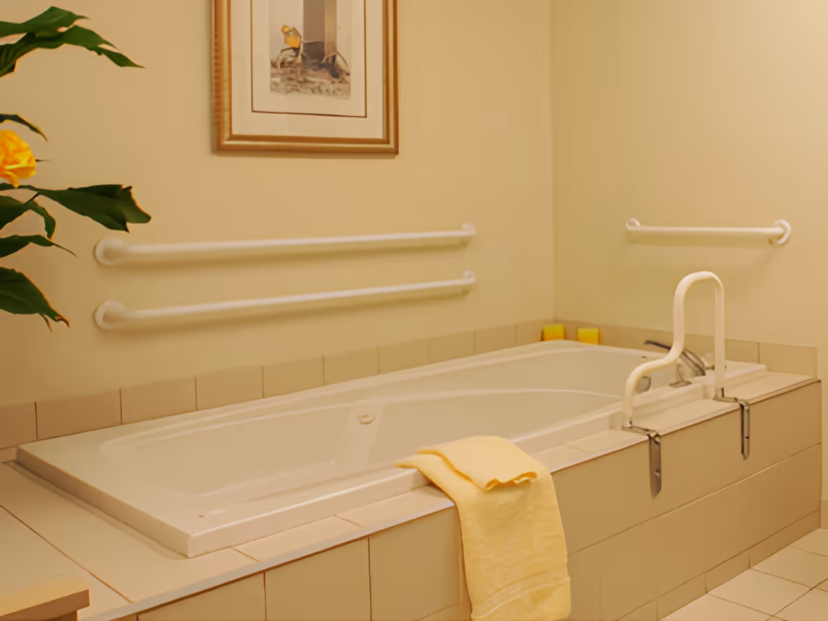 Accessible built-in bathtub with grab bars, a yellow towel draped over the edge, and framed artwork on a pale wall.