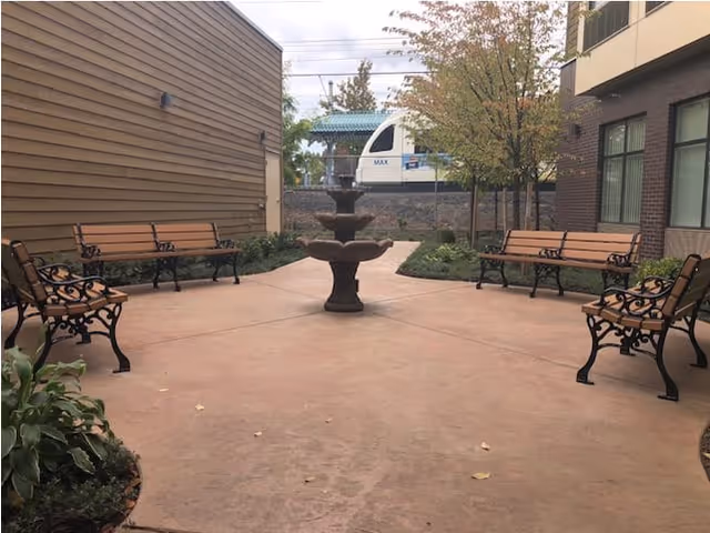 Outdoor courtyard area with a central three-tiered fountain surrounded by six wooden benches with black metal frames. The courtyard is paved and bordered by plants and trees, with a building on the right and a wooden fence on the left. A train is visible in the background beyond the fence.