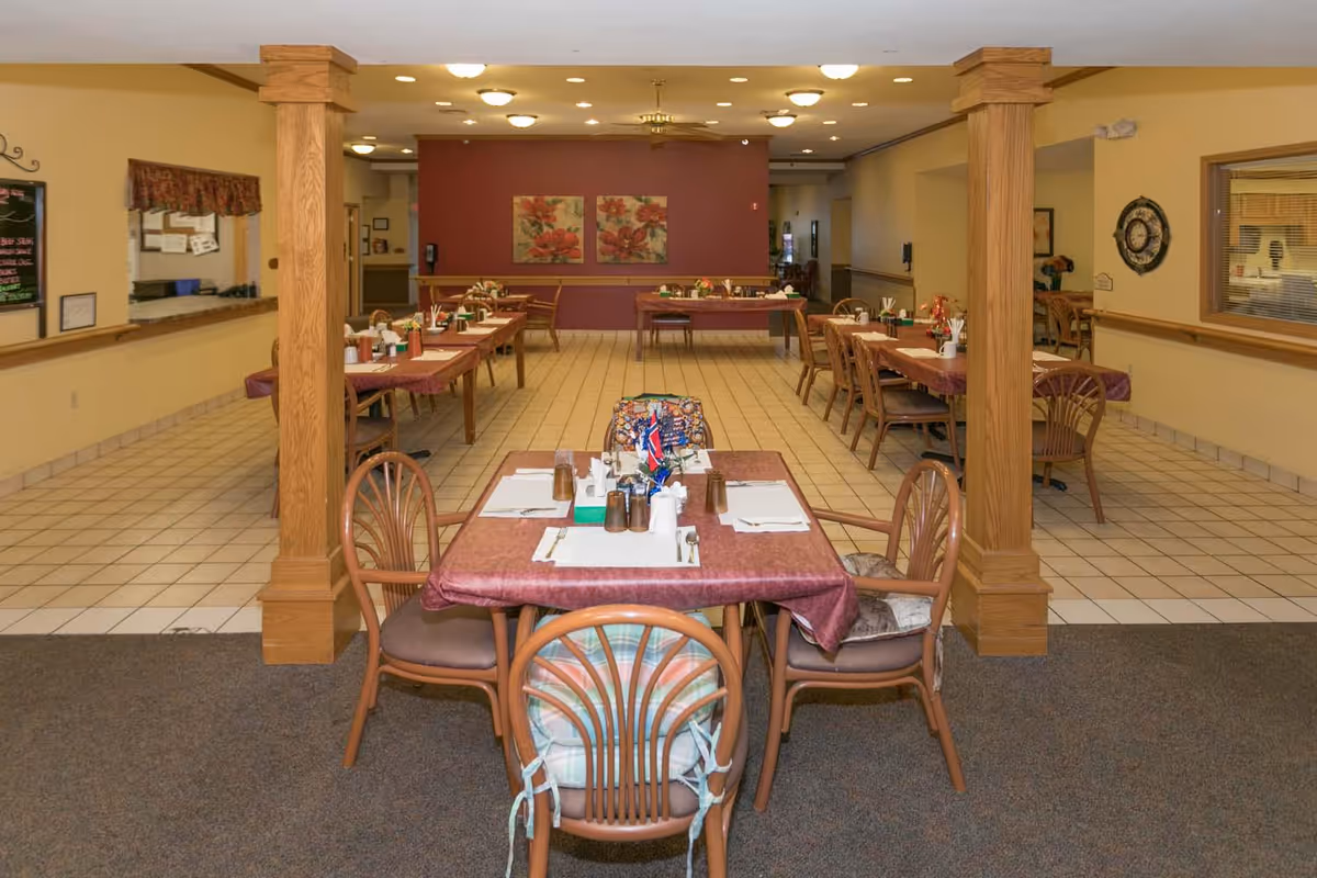 Communal dining room with several tables set for meals, chairs, and wooden support columns.