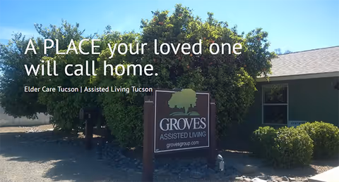 Exterior view of Groves Assisted Living facility with a sign in front displaying the facility name and website. The building is surrounded by green bushes and trees under a clear blue sky.