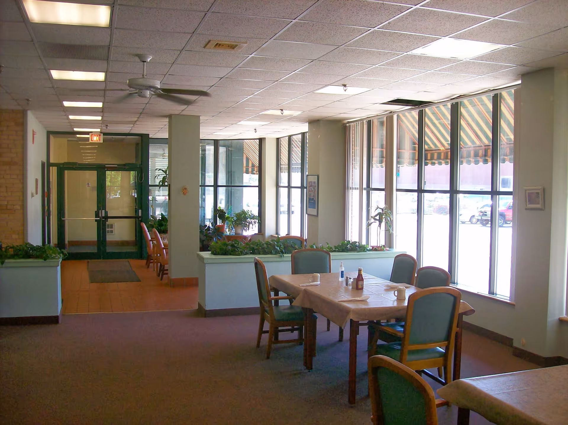 Interior view of a senior living facility dining area with tables and chairs. Large windows with striped awnings outside let in natural light. There are plants on ledges and a ceiling fan above. The entrance door is visible in the background.