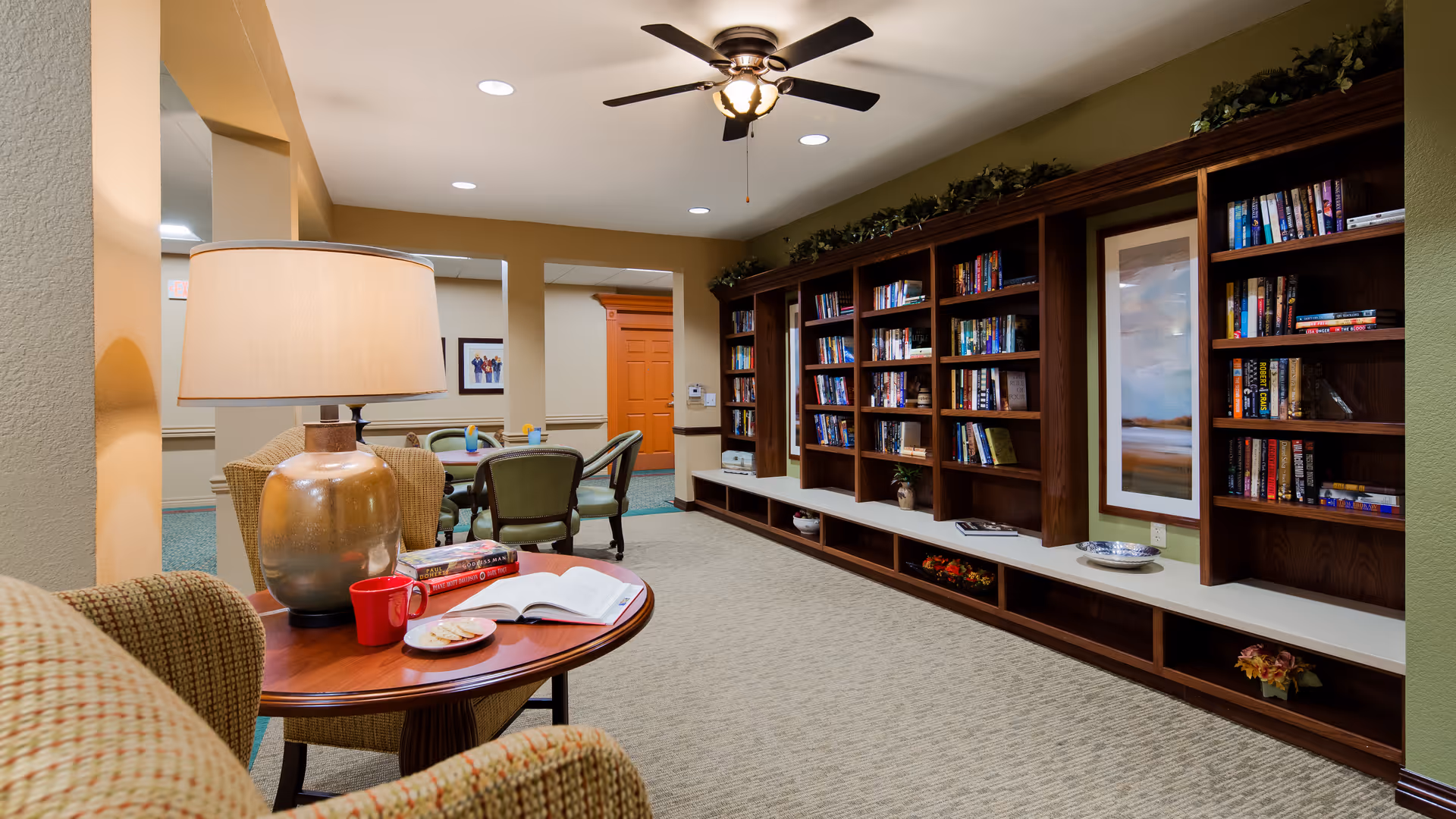 A cozy communal library-style lounge with built-in bookshelves, seating, a side table with a lamp and open book, and a ceiling fan.